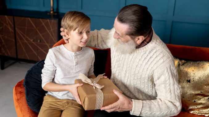 Moment touchant entre générations lors d'un échange de cadeau Enfant blond offrant cadeau emballé à grand-père barbu sur canapé, moment d'offrir un cadeau à son père